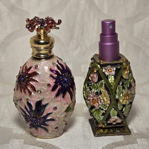 May include: Two decorative perfume bottles. One is pink with floral designs and jewel accents, topped with a gold cap and a red floral ornament. The other is a square-based bottle with a floral pattern and a purple spray top.