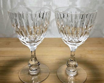 Puede incluir: Dos copas de vino de cristal transparente con intrincados patrones tallados. Las copas tienen un cuenco en forma de tulipán, un tallo delgado con un nudo decorativo y una base redonda. Se muestran sobre una superficie de madera.