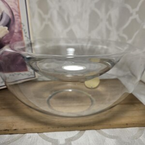 May include: Two clear glass bowls, one nested inside the other. The larger bowl sits on a wooden surface, and the smaller bowl is suspended within it. The bowls are transparent, allowing light to pass through. The background is a white patterned fabric.