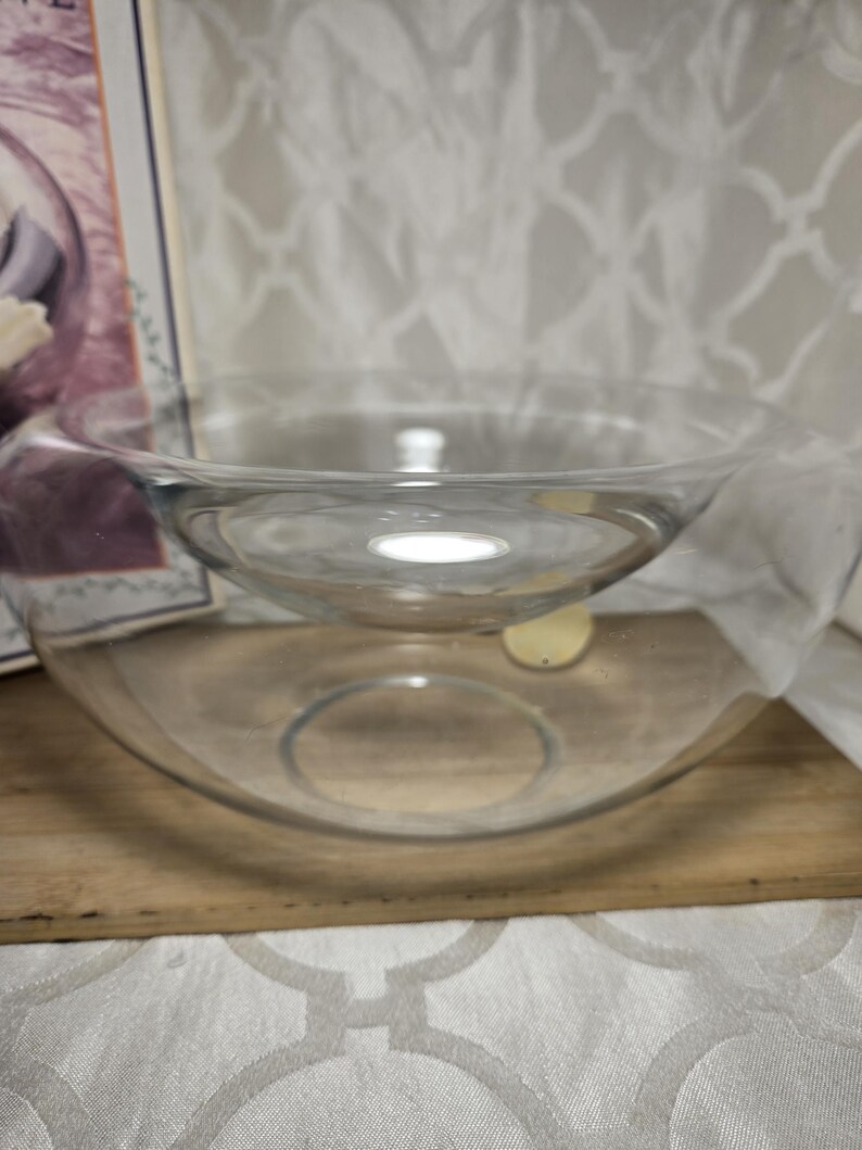 May include: Two clear glass bowls, one nested inside the other. The larger bowl sits on a wooden surface, and the smaller bowl is suspended within it. The bowls are transparent, allowing light to pass through. The background is a white patterned fabric.