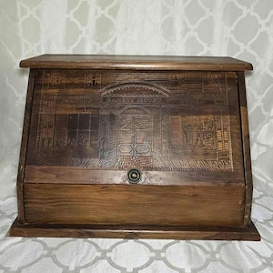 May include: A dark brown wooden bread box with a slanted front door featuring an engraved illustration of a storefront labeled BAKERY HOUSE. The box has a rectangular base, a flat top, and a small circular metal knob centered on the front panel.