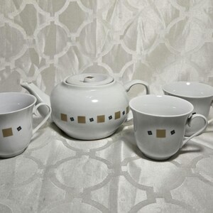 May include: A white ceramic tea set with a teapot and three cups. The teapot and cups are decorated with a repeating pattern of gold squares and black diamonds. The set is displayed on a white patterned surface.