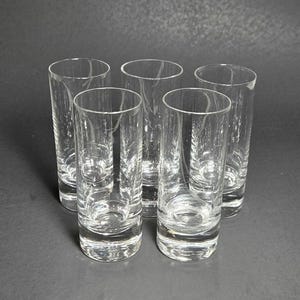 May include: Five clear, cylindrical drinking glasses. The glasses are made of transparent glass, allowing a view of the contents. The glasses have a simple, elegant design, suitable for various beverages. The glasses are arranged in a circular pattern.