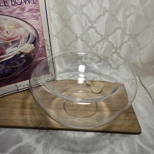 May include: A clear glass bowl with a wide opening, resting on a wooden surface. The bowl is circular and appears to be empty. A small, light-colored object sits inside the bowl. The packaging for the bowl is visible in the background.