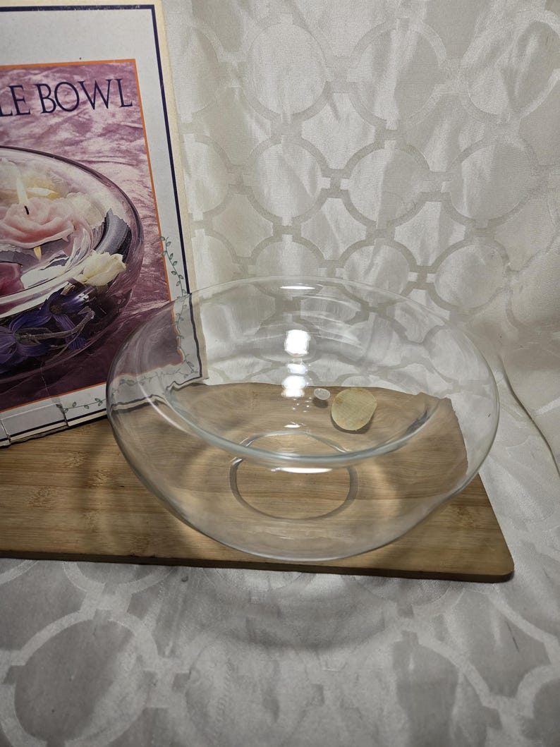 May include: A clear glass bowl with a wide opening, resting on a wooden surface. The bowl is circular and appears to be empty. A small, light-colored object sits inside the bowl. The packaging for the bowl is visible in the background.