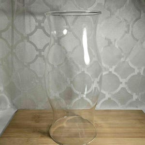 May include: A clear glass hurricane candle holder with a flared top and a rounded base. The candle holder is sitting on a wooden cutting board. The background is a patterned, light-colored fabric.