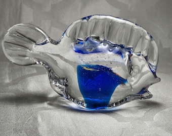 May include: A clear glass sculpture in the shape of a fish. The sculpture has a vibrant blue section in the center. The fish has a tail and fins, and the glass is textured. The sculpture is on a white surface.