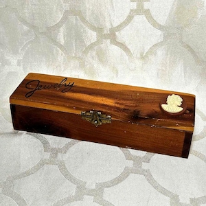 May include: Rectangular wooden jewelry box with a hinged lid. The box is stained a warm brown color and features the word "Jewelry" in a cursive font. A cameo is affixed to the right side. The box has a brass clasp.