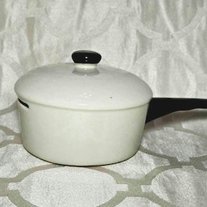 May include: A small, white ceramic saucepan with a lid and a black handle. The lid has a black knob. The saucepan is on a patterned white surface. This kitchenware item is suitable for cooking small portions.