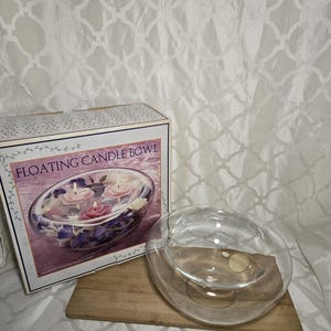 May include: A clear glass floating candle bowl with a wooden base, next to a box with a picture of the bowl with floating candles and flowers. The box reads "FLOATING CANDLE BOWL". The background is a white patterned fabric.
