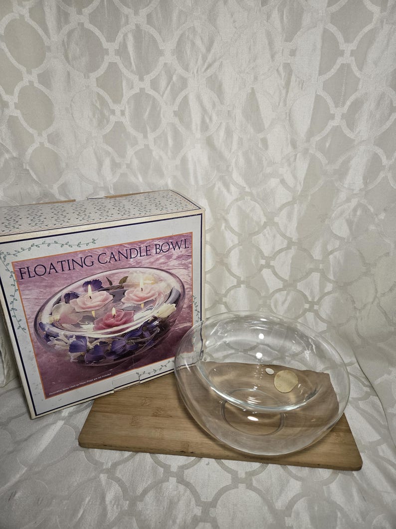 May include: A clear glass floating candle bowl with a wooden base, next to a box with a picture of the bowl with floating candles and flowers. The box reads "FLOATING CANDLE BOWL". The background is a white patterned fabric.