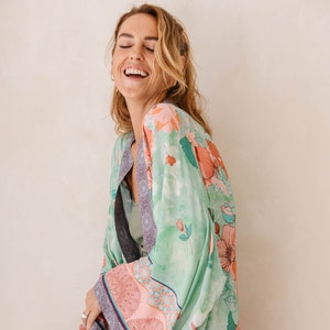 May include: A woman wearing a green floral kimono with pink and blue accents. The kimono has a long, flowing silhouette and is made of a lightweight fabric.