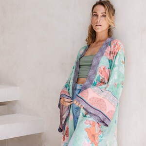 May include: A woman wearing a long, flowing green and pink floral kimono with a purple trim. The kimono has a pink and white patterned border. The woman is standing in front of a white wall.
