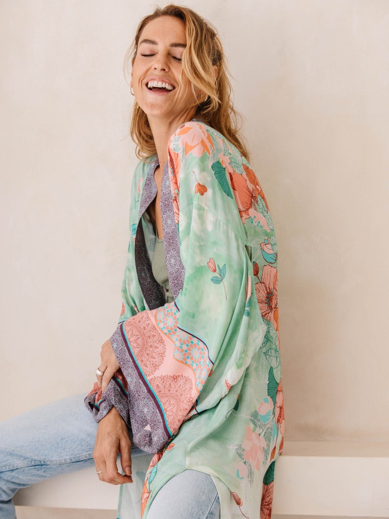 May include: A woman wearing a green floral kimono with pink and blue accents. The kimono has a long, flowing silhouette and is made of a lightweight fabric.
