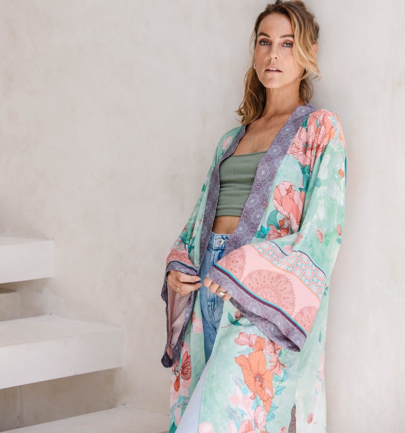 May include: A woman wearing a long, flowing green and pink floral kimono with a purple trim. The kimono has a pink and white patterned border. The woman is standing in front of a white wall.
