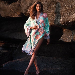 May include: A woman wearing a floral patterned kimono with a light green and pink color scheme. The kimono has a long, flowing design and is worn over a white tank top. The woman is standing in front of a rocky background.