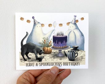 Può includere: Illustrazione di biglietto di compleanno con due figure di fantasmi con cappellini da festa, un gatto nero e una torta viola con candele. La carta ha il testo "Have a Spooktacular Birthday!" ed è decorata con zucche.