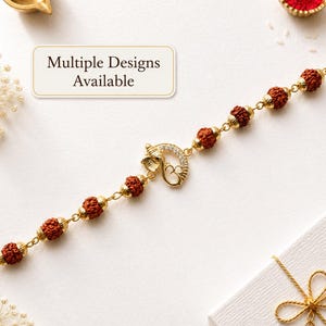 Op de afbeelding: Een armband met bruine rudraksha-kralen afgewisseld met goudkleurige metalen tussenstukjes en een centrale goudkleurige bedel met een Om-symbool en een Ganesha-figuur versierd met kleine heldere steentjes. De tekst Multiple Designs Available is zichtbaar.