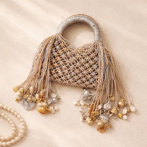 Puede incluir: Un bolso tejido con un asa circular y un cuerpo en forma de red. El bolso está adornado con largas borlas y elementos decorativos, incluyendo conchas y cuentas. La paleta de colores incluye tonos dorados, plateados y beige, creando una estética bohemia.