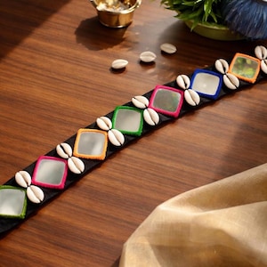 May include: A black belt featuring square mirrors with colorful borders and white seashells. The mirrors are framed in orange, green, pink, and blue. The belt is displayed on a wooden surface, with a gold bowl and greenery in the background.