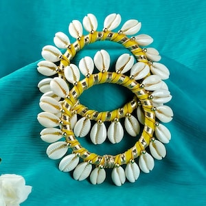 Może przedstawiać: Dwa żółte i złote bransoletki z przymocowanymi białymi muszelkami. Bransoletki leżą na turkusowym tle.