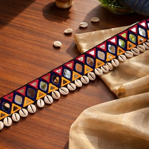 May include: A decorative trim featuring a black base with colorful geometric patterns in red, yellow, and blue, interspersed with small mirrors. The trim is adorned with a row of white seashells along one edge, creating a unique and textured design.
