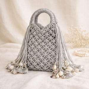 Puede incluir: Un bolso de mano de macramé gris con un asa redondeada y largas borlas. El bolso presenta un patrón tejido y está adornado con borlas decorativas, cuentas y conchas. Un accesorio elegante.