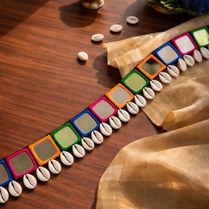 Puede incluir: Un cinturón colorido con detalles de espejos cuadrados en varios colores, incluyendo rosa, verde y azul, con conchas blancas a lo largo del borde inferior. El cinturón se muestra sobre una superficie de madera con un cuenco dorado y tela en el fondo.