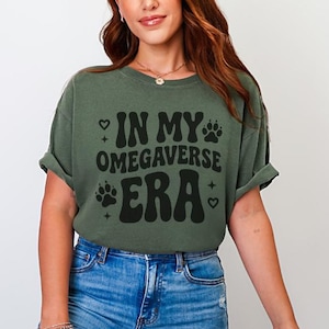 May include: Olive green t-shirt with the text "IN MY OMEGAVERSE ERA" in black. The design includes paw prints and hearts. The shirt is paired with blue jeans.