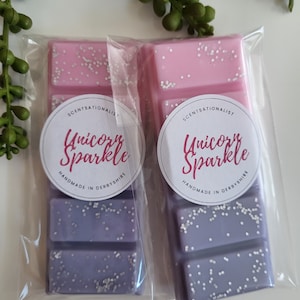 Può includere: Due cere per profumare l'ambiente in una confezione trasparente. Le cere sono stratificate in rosa e viola con glitter bianco. La confezione ha un'etichetta circolare bianca con il testo "ScentSationalist Unicorn Sparkle Handmade in Derbyshire".