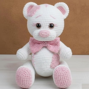 Peut inclure: Un ours en peluche crocheté blanc fait à la main avec des détails roses. L'ours porte un nœud papillon rose, des taches roses et des détails roses sur les oreilles et les pieds. Il a de grands yeux bruns et un nez rose.