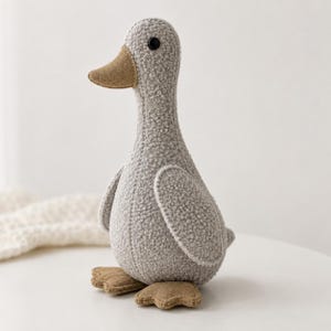 Peut inclure: Un canard en peluche avec un corps texturé gris clair en tissu bouclé. Le canard possède un bec lisse de couleur beige et des pattes palmées assorties. Un petit œil circulaire noir est placé sur le côté de la tête, et le corps présente des coutures visibles le long du cou et des ailes.