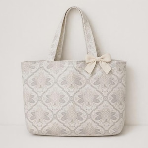 May include: A tote bag featuring a repeating light gray and cream damask-style pattern. The bag has two top handles and a small, solid cream-colored bow attached to the upper right side of the front panel, providing a decorative accent.