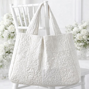 Könnte beinhalten: Eine weiße, gesteppte Tragetasche mit floralen Mustern und zwei Griffen. Die Tasche besteht aus einem strukturierten Stoff und steht auf einem weißen Stuhl. Der Hintergrund umfasst weiße Blumen.