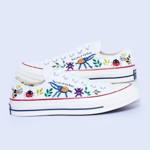Op de afbeelding: Een paar witte lage canvas sneakers met handgeschilderde kleurrijke insecten, groene bladeren en de tekst "The Lost of the Bugs" op de zijkant. De schoenen hebben witte veters, een witte rubberen neus en een rode en blauwe streep op de zool.