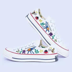 Op de afbeelding: Een paar witte canvas sneakers met kleurrijke illustraties van diverse insecten, waaronder kevers, lieveheersbeestjes en bijen, samen met groen blad. De tekst "The Lost of the Bugs" staat gedrukt op de zijkant van elke schoen boven de rubberen zool.