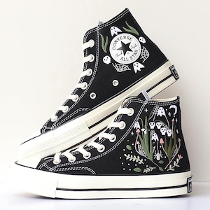 May include: Pair of black high-top Converse Chuck Taylor All Star sneakers with white laces and soles. The sneakers feature embroidered Halloween-themed designs, including ghosts, spiderwebs, and the words "Converse All Star".