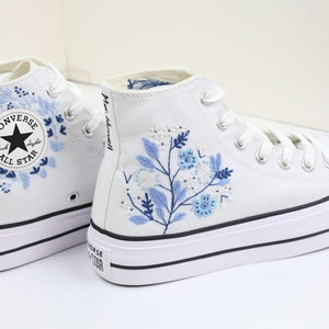 May include: White high-top Converse sneakers with blue floral embroidery. The Converse All Star logo is visible on the side of the shoe.