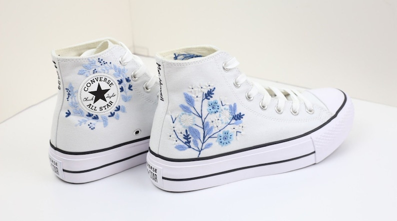 May include: White high-top Converse sneakers with blue floral embroidery. The Converse All Star logo is visible on the side of the shoe.