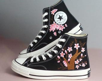 May include: Black high-top sneakers with white laces and soles. The sneakers feature a floral design with pink blossoms and brown branches. The Converse All Star logo is visible on the side.