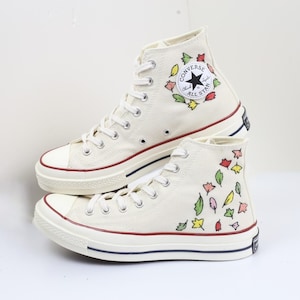 May include: A pair of white high-top Converse sneakers with a floral design. The sneakers have a red trim around the sole and a black Converse All Star logo on the side.
