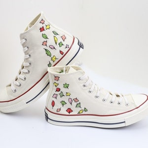 K&ouml;nnte beinhalten: Wei&szlig;e High-Top-Canvas-Sneaker mit einem roten, gelben, gr&uuml;nen und pinkfarbenen Blatt-Muster, das an die Seite gemalt ist.