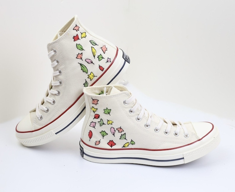 K&ouml;nnte beinhalten: Wei&szlig;e High-Top-Canvas-Sneaker mit einem roten, gelben, gr&uuml;nen und pinkfarbenen Blatt-Muster, das an die Seite gemalt ist.