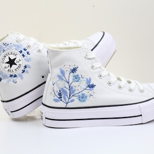 May include: White high-top Converse sneakers with a platform sole. The sneakers feature blue floral embroidery and a black Converse All Star logo.