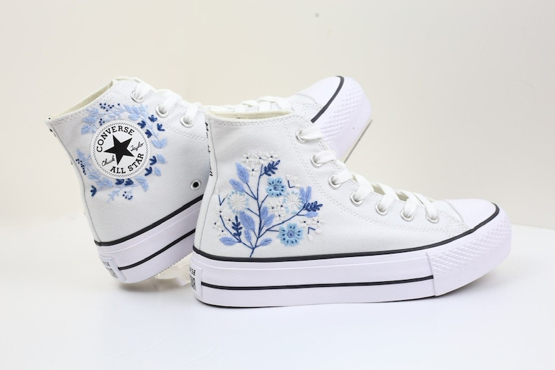 May include: White high-top Converse sneakers with a platform sole. The sneakers feature blue floral embroidery and a black Converse All Star logo.