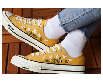 May include: Yellow canvas sneakers with white laces and embroidered white flowers. The shoes have a white rubber sole and the text "HANA STORE" is printed on the side.
