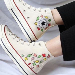 K&ouml;nnte beinhalten: Wei&szlig;e Converse High-Top-Sneaker mit rotem und blauem Besatz. Die Sneaker sind mit handbemalten bunten Bl&auml;ttern verziert. Das Converse-Logo ist an der Seite des Schuhs sichtbar.