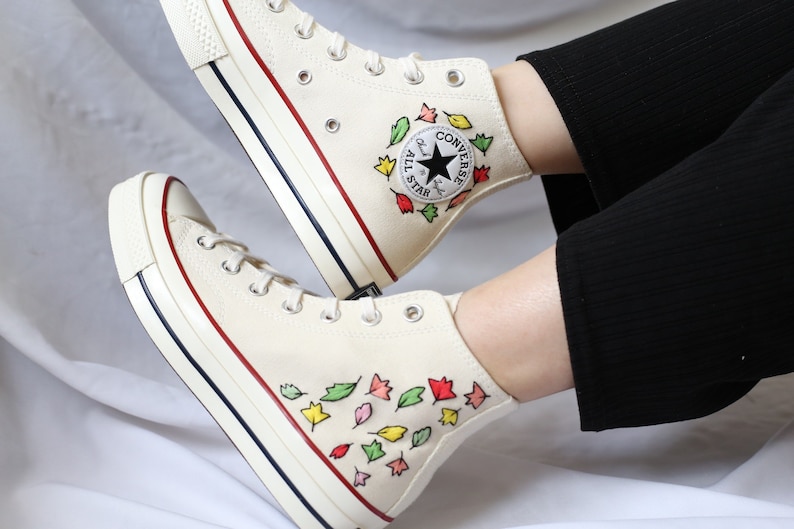 K&ouml;nnte beinhalten: Wei&szlig;e Converse High-Top-Sneaker mit rotem und blauem Besatz. Die Sneaker sind mit handbemalten bunten Bl&auml;ttern verziert. Das Converse-Logo ist an der Seite des Schuhs sichtbar.