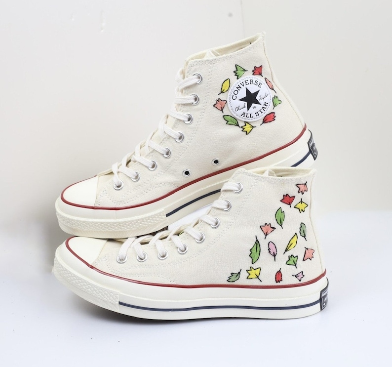 K&ouml;nnte beinhalten: Ein Paar wei&szlig;e Converse-Sneaker mit hohem Schaft und floralem Muster. Die Sneaker haben einen roten Rand um die Sohle und ein schwarzes Converse All Star Logo an der Seite.