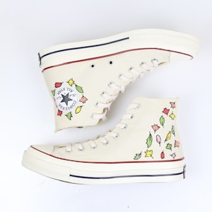 K&ouml;nnte beinhalten: Wei&szlig;e Converse-Sneaker mit hohem Schaft, floralem Muster und schwarzem Stern-Logo. Der Schriftzug "Converse All Star" ist an der Seite des Schuhs aufgedruckt.